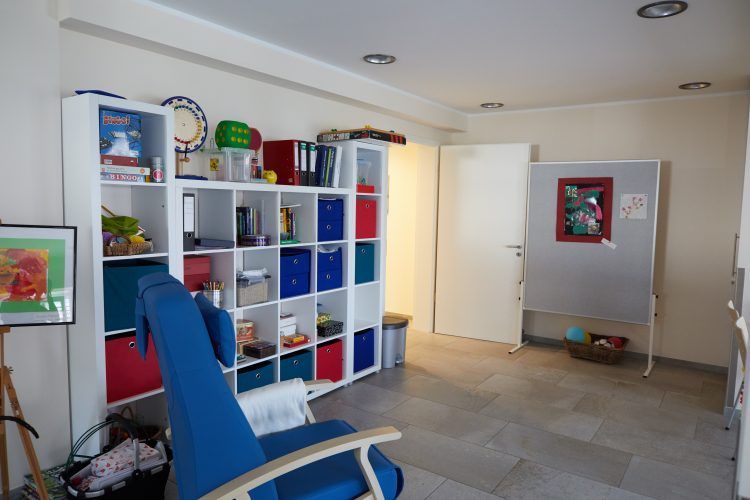Helles Zimmer mit Regalen, blauem Stuhl, offener Tür und kleinem Wandspiegel