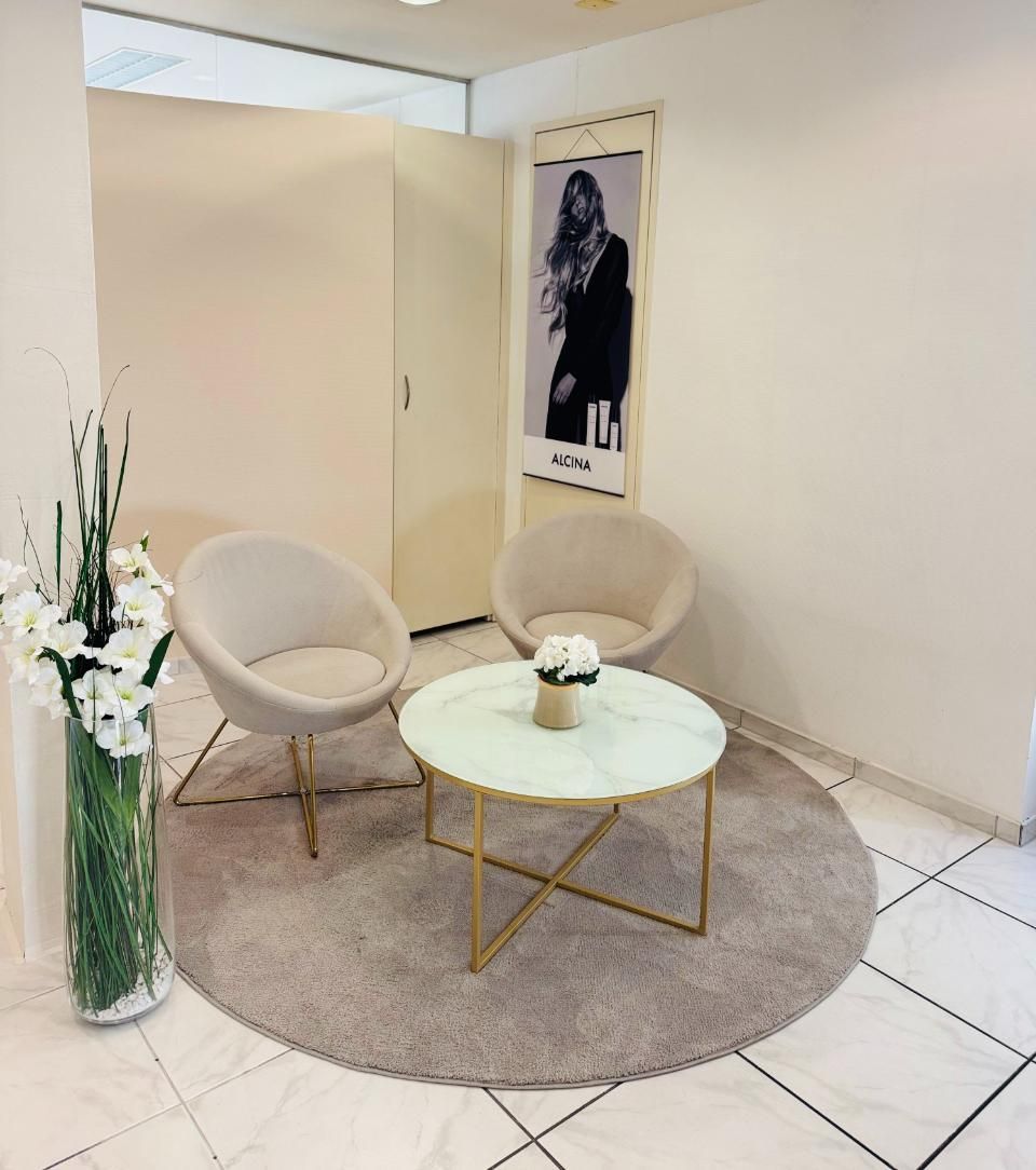 Kleine Lounge-Ecke mit zwei beigen Stühlen, rundem Tisch, grauem Teppich und Wandbildern auf weissem Fliesenboden.