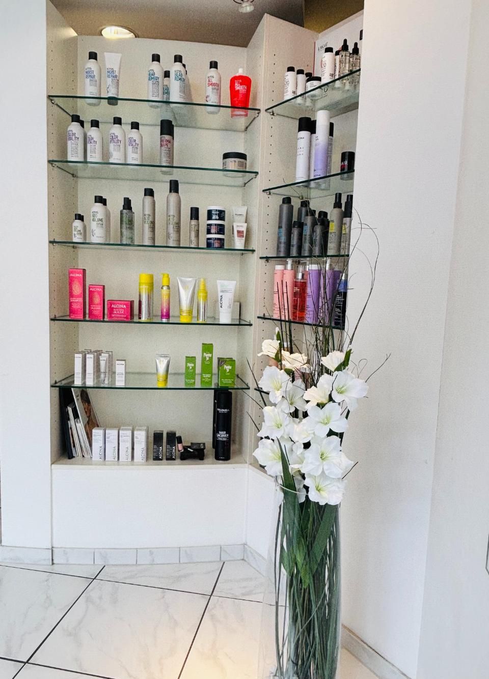 Regale voller Schönheitsprodukte in einem hellen Salon, an der Wand eine Vase mit weissen Blumen.