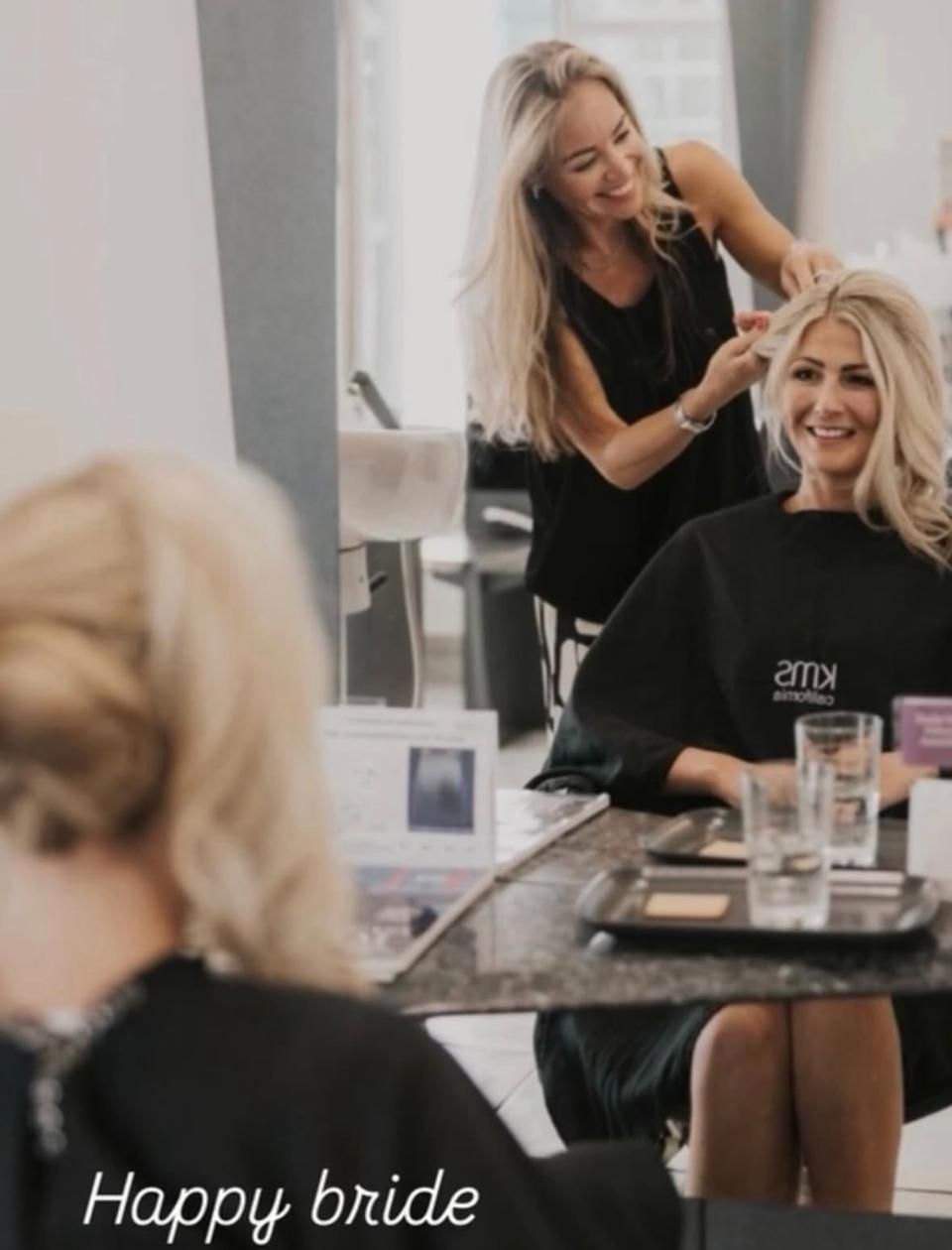 Eine Braut lächelt auf einem Friseurstuhl, während ihr eine Stylistin die blonden Haare macht.