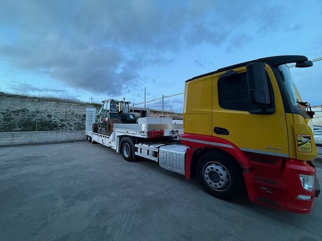 Camión amarillo y rojo con remolque de plataforma que transporta equipos de construcción; entorno al aire libre.