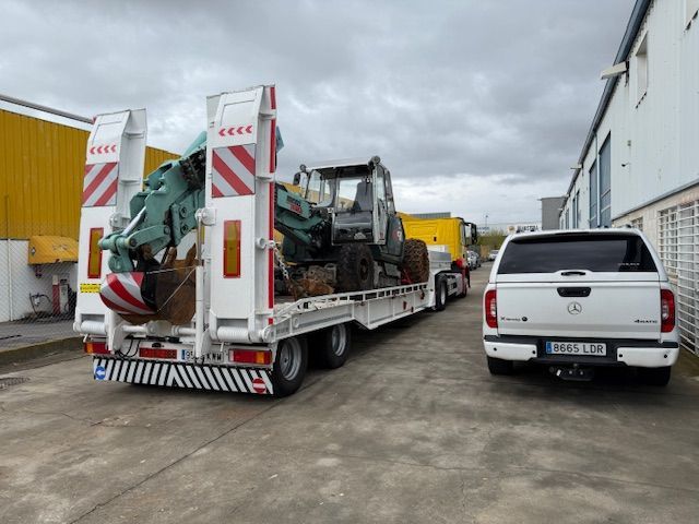 Un gran remolque blanco transporta un vehículo de construcción con una camioneta blanca a su lado.