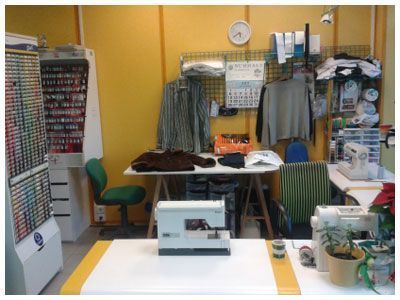 Pequeño taller o cuarto de trabajo desordenado, con estanterías, ropa colgada, mesas, sillas y materiales, junto a paredes amarillas.