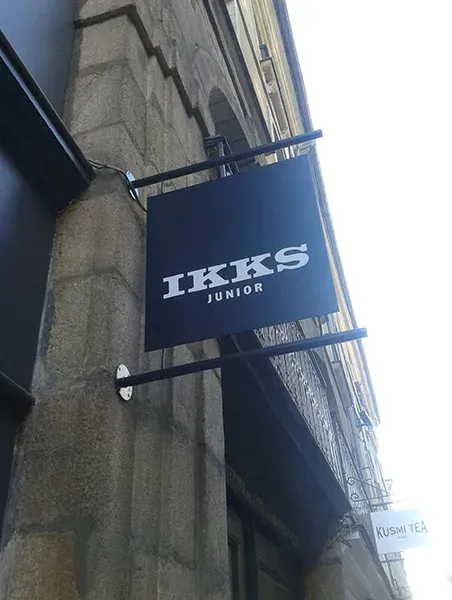 Enseigne noire IKKS suspendue sur la façade en pierre d’un bâtiment.