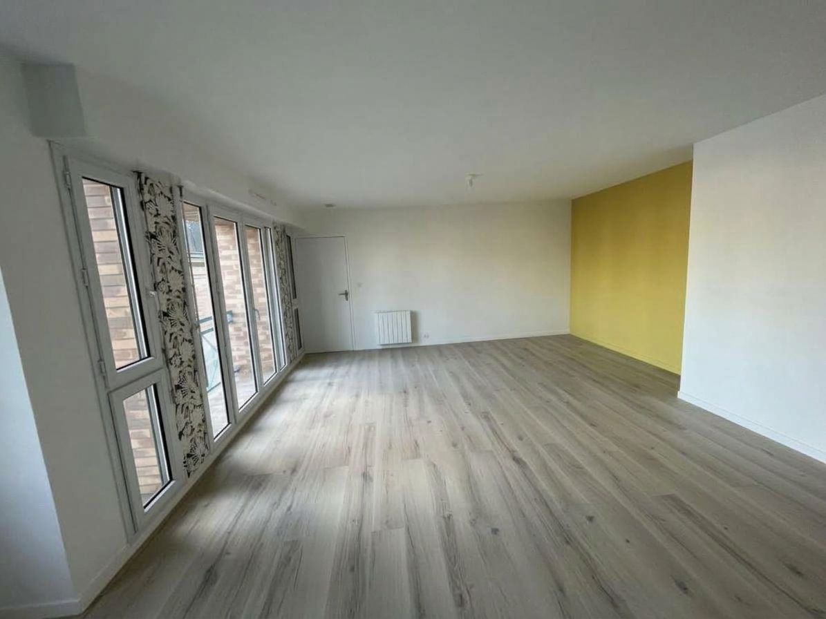 Pièce moderne vide avec parquet, grandes fenêtres et mur d’accent jaune.