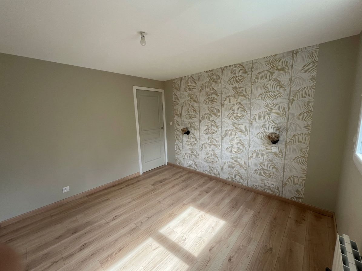 Pièce vide avec un parquet clair, un mur d’accent au papier peint fleuri et la lumière du soleil venant d’une fenêtre.