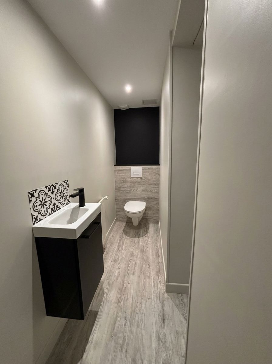 Toilettes modernes et étroites avec lavabo et WC, murs gris, sol carrelé et mur d’accent foncé.