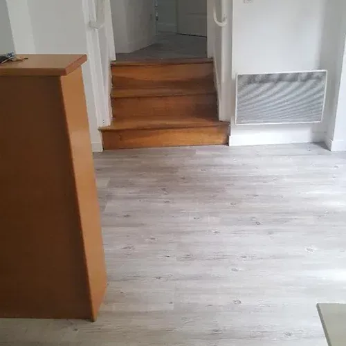 Pièce vide avec un parquet clair, des marches en bois et une bouche d’aération murale près d’une porte.