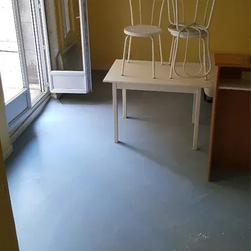Petite pièce avec un sol bleu, une table blanche et des tabourets près d’une porte vitrée.