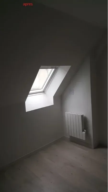 Pièce mansardée vide avec un plafond en pente, une fenêtre de toit et un radiateur blanc sur un mur gris