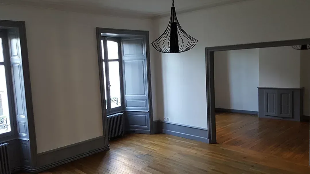 Pièce vide avec parquet, murs blancs, moulures foncées et luminaire suspendu.