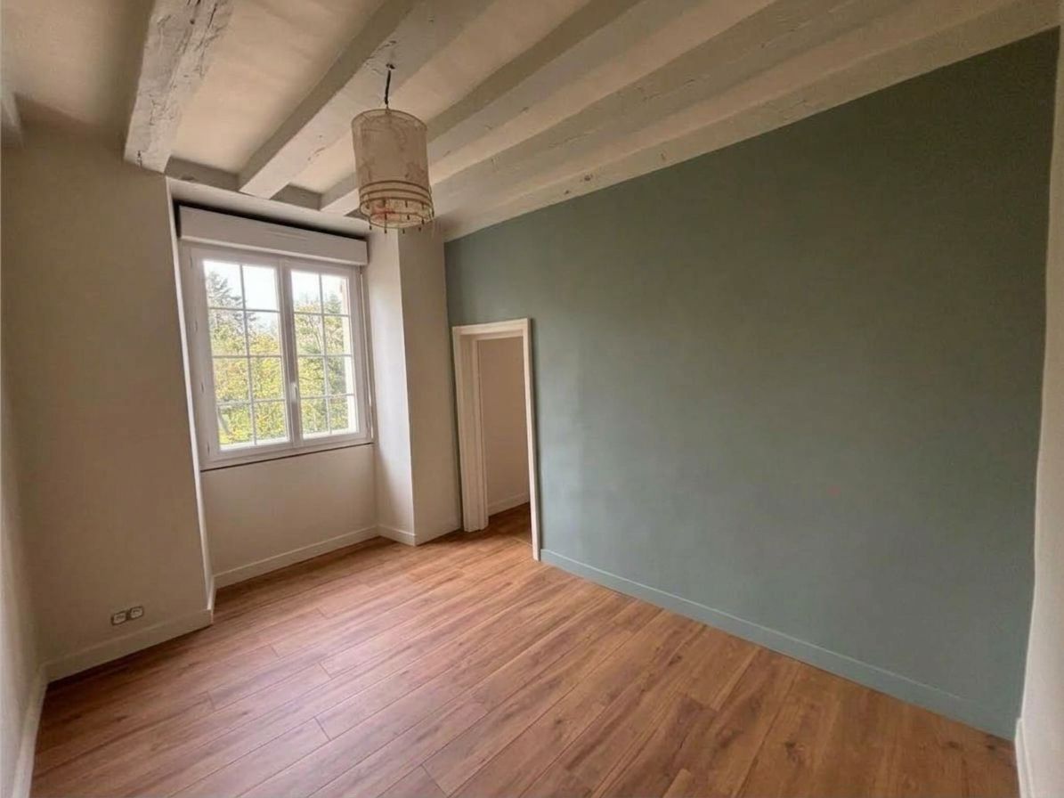 Pièce vide avec un parquet, un mur vert, des poutres apparentes et une fenêtre laissant entrer la lumière du jour.