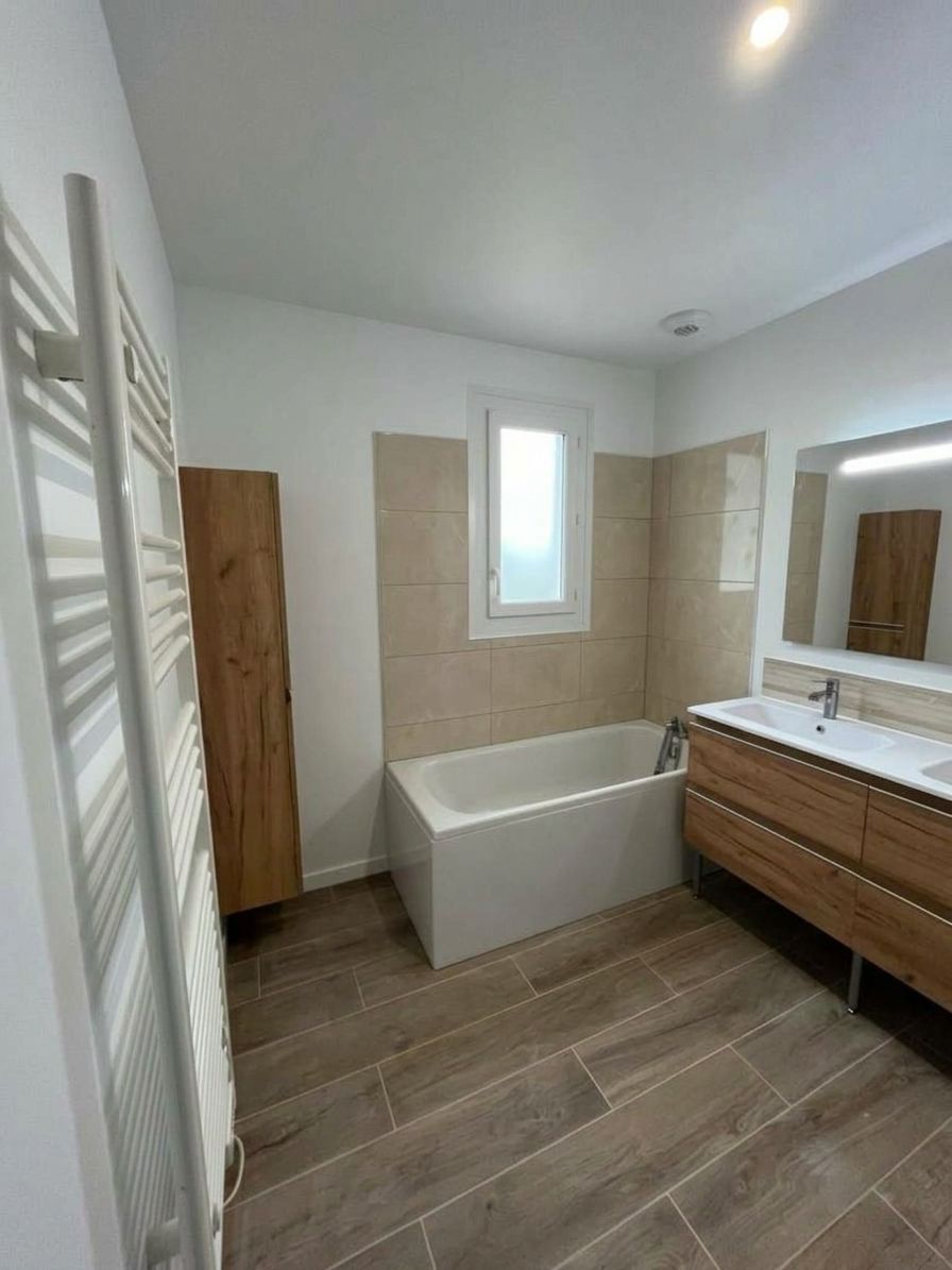 Salle de bains moderne avec baignoire, murs carrelés beige, meuble vasque en bois et radiateur sèche-serviettes blanc