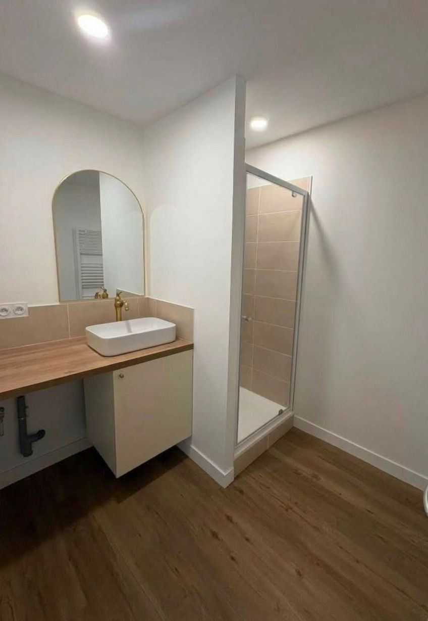 Petit meuble-lavabo avec miroir et vasque, à côté d’une douche ouverte, murs clairs et sol imitation bois.
