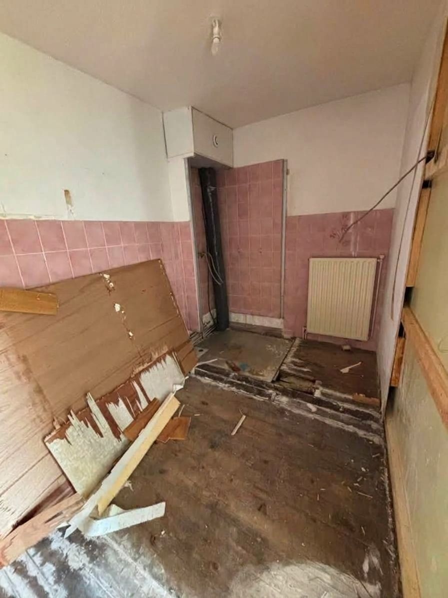 Pièce endommagée avec un sol défoncé, des murs carrelés en rose et une porte au fond.