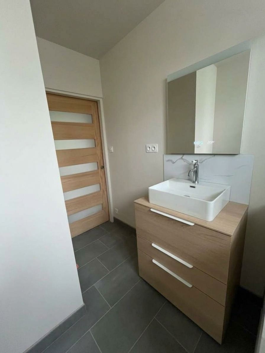 Salle de bains moderne avec meuble-vasque en bois, lavabo blanc, miroir et porte en verre dépoli.