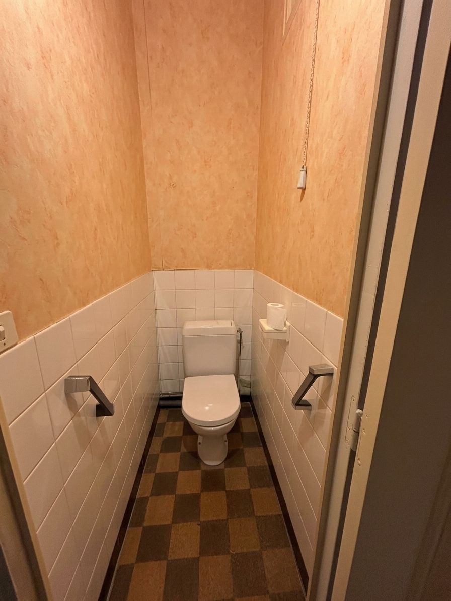 Cabine de toilettes étroite avec des WC, des carreaux blancs, des murs beiges et un sol à carreaux