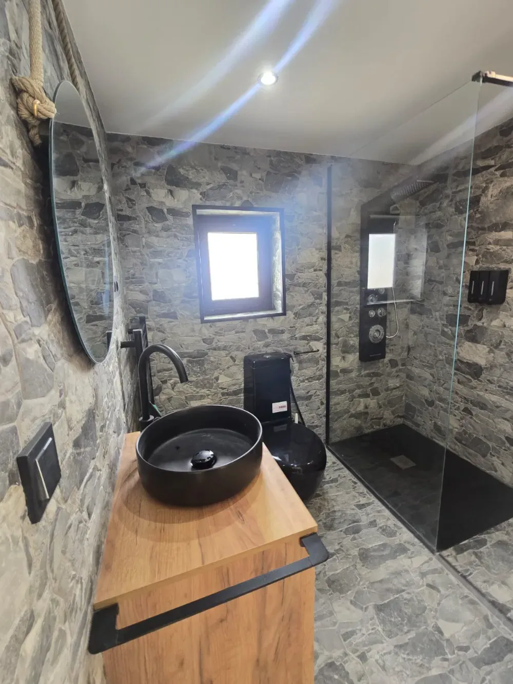 Baño con paredes de piedra, accesorios negros, tocador de madera, espejo redondo y ducha de cristal.