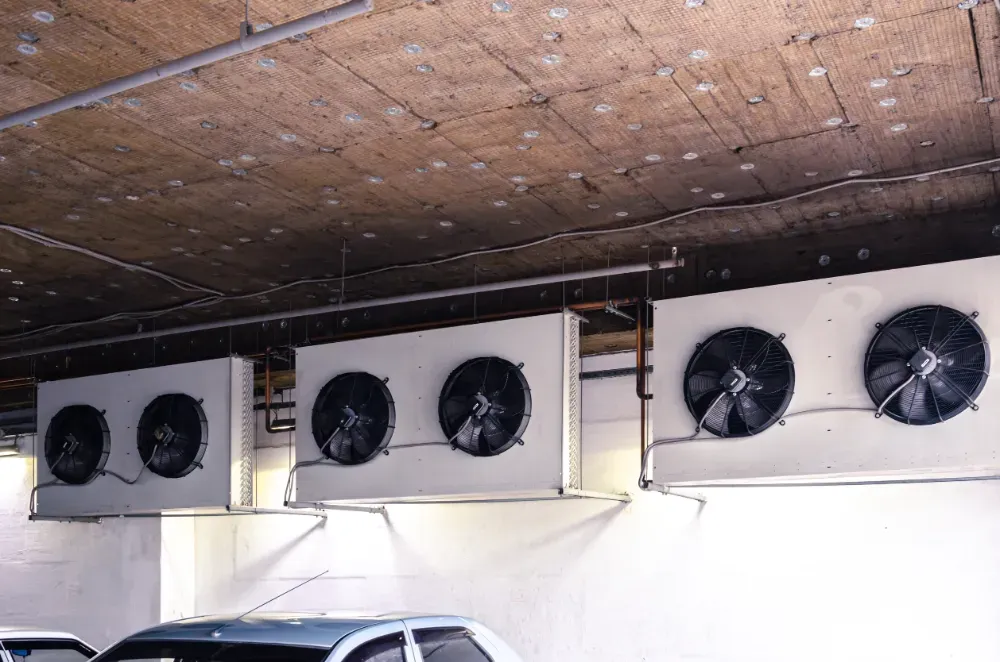 Cuatro grandes unidades de aire acondicionado blancas suspendidas de un techo de hormigón en un estacionamiento.