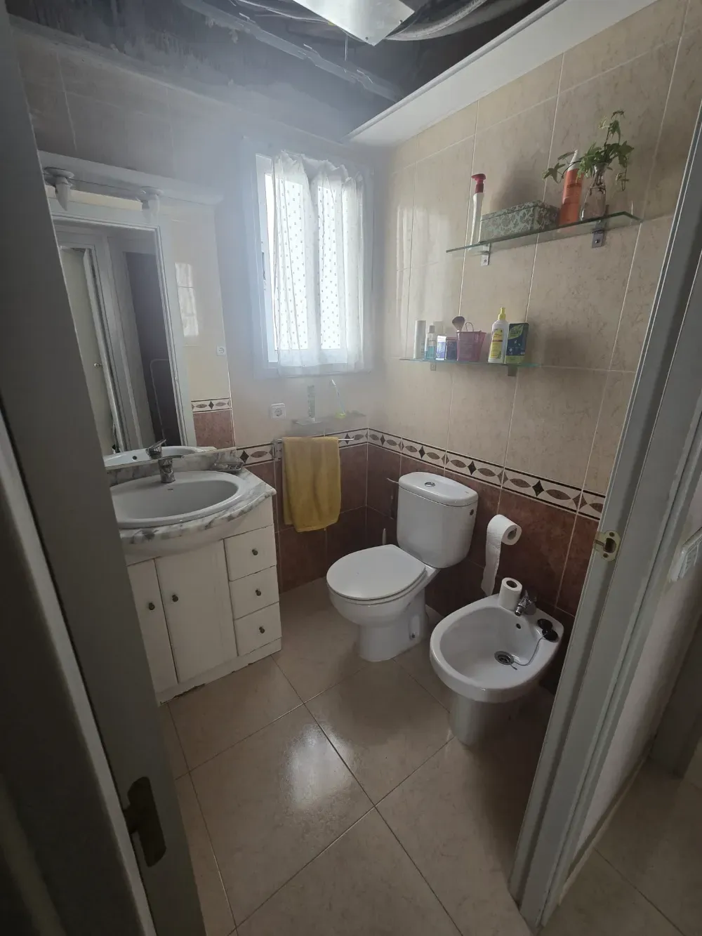 Baño con inodoro, bidé, lavabo y ventana. Azulejos beige y marrón. Toalla amarilla.