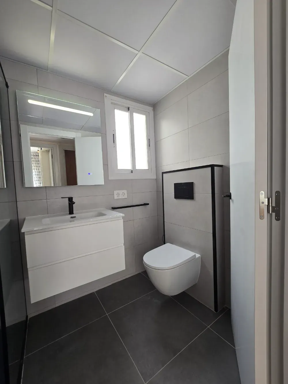 Baño moderno con lavabo blanco, inodoro flotante y azulejos grises.