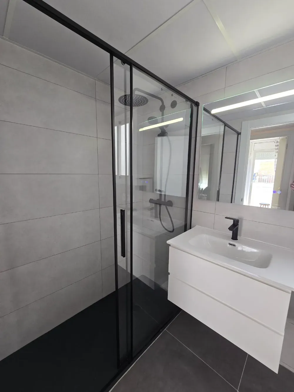 Baño moderno con ducha de cristal, tocador y espejo grande. Combinación de colores gris y negro.