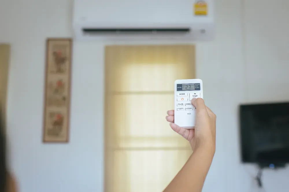 Mano sosteniendo el control remoto del aire acondicionado y apuntándolo hacia la unidad montada en la pared.