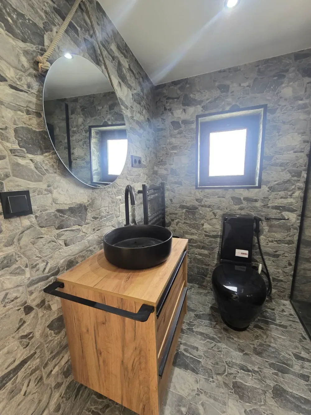 Baño con paredes de imitación piedra, tocador de madera con lavabo negro e inodoro negro. Un gran espejo redondo cuelga.