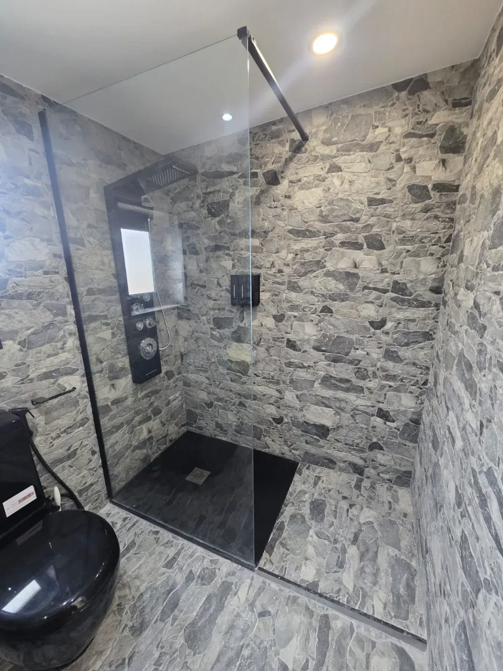 Baño con pared y suelo con motivos de piedra, cabina de ducha de color negro y inodoro de color negro.