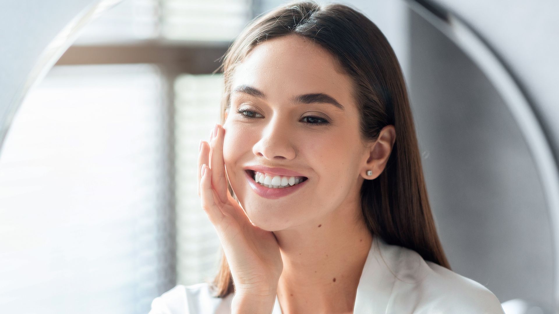 Femme souriante, se touchant la joue, se regardant dans un miroir. Blazer, fond neutre.