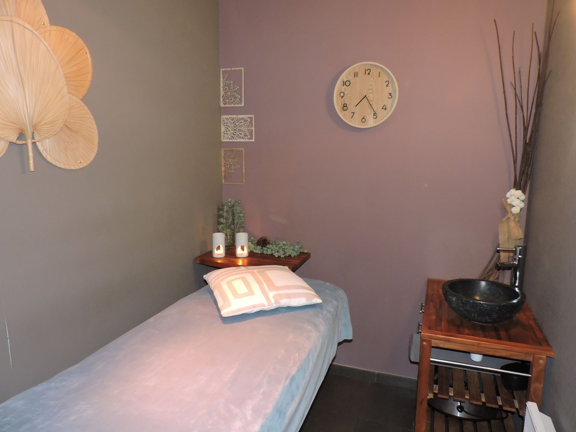 Salle de massage aux murs, table de massage, étagère en bois avec un vase, horloge et ventilateur décoratif.