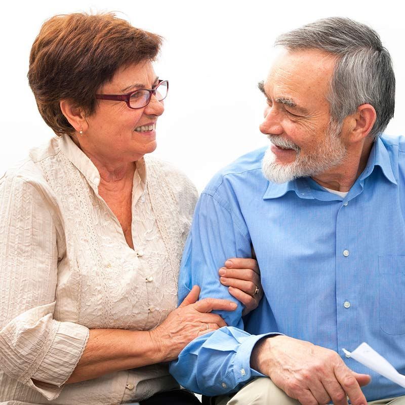 Una pareja sonriente sentada muy cerca la una de la otra, tomadas de la mano y mirándose el uno al otro.