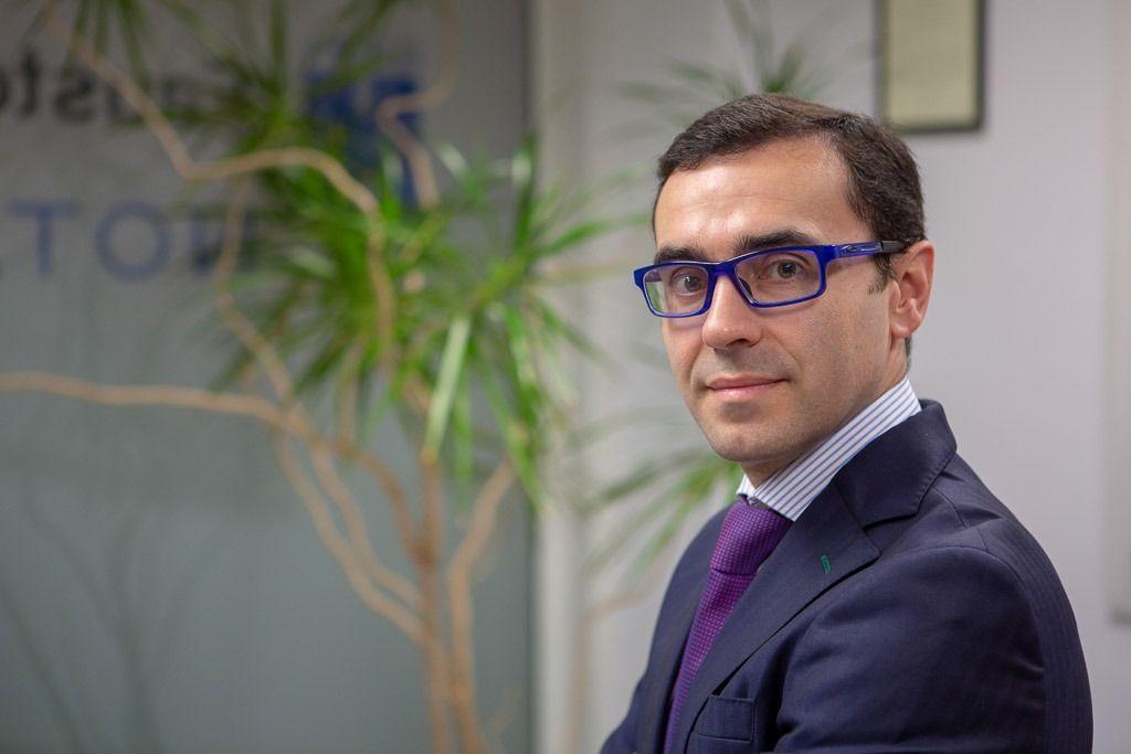 Hombre con traje azul y gafas de pie en una oficina, mirando a la cámara.