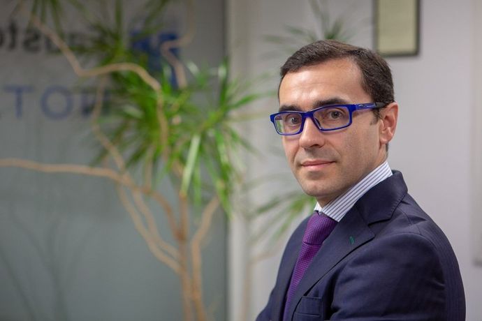 Hombre con traje azul y gafas de pie en una oficina, mirando a la cámara.