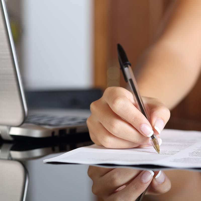 Una mano sostiene un bolígrafo sobre un papel junto a una computadora portátil en un escritorio.