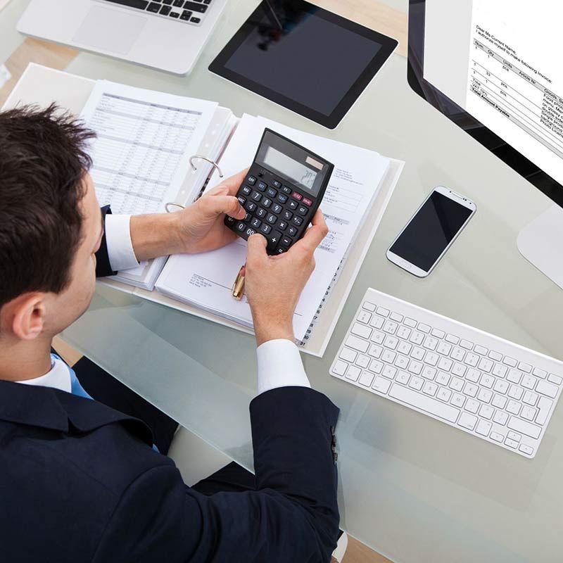 Persona utilizando una calculadora en un escritorio con papeles, ordenador portátil, teclado y teléfono inteligente en una oficina luminosa.