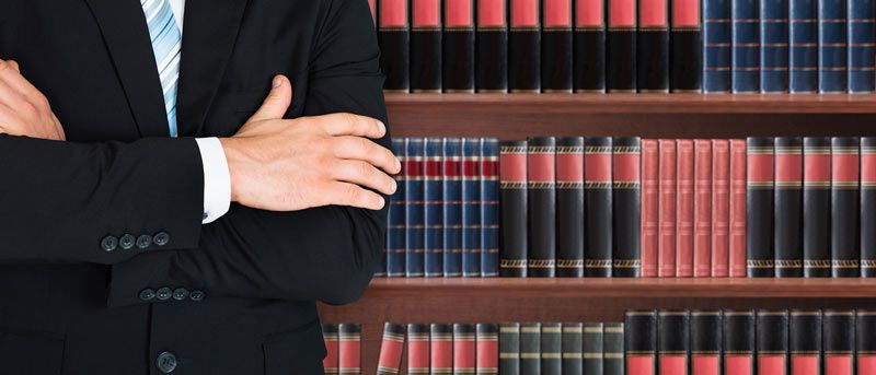 Hombre de negocios con traje oscuro y brazos cruzados frente a libros de leyes rojos y negros en estanterías.