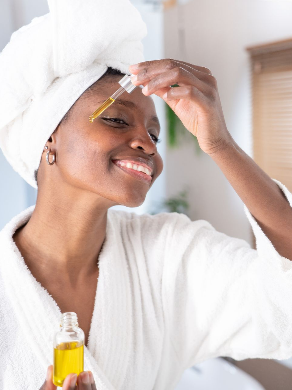 Une personne souriante, vêtue d'un peignoir et d'une serviette blancs, applique un sérum facial jaune à l'aide d'un compte-gouttes.