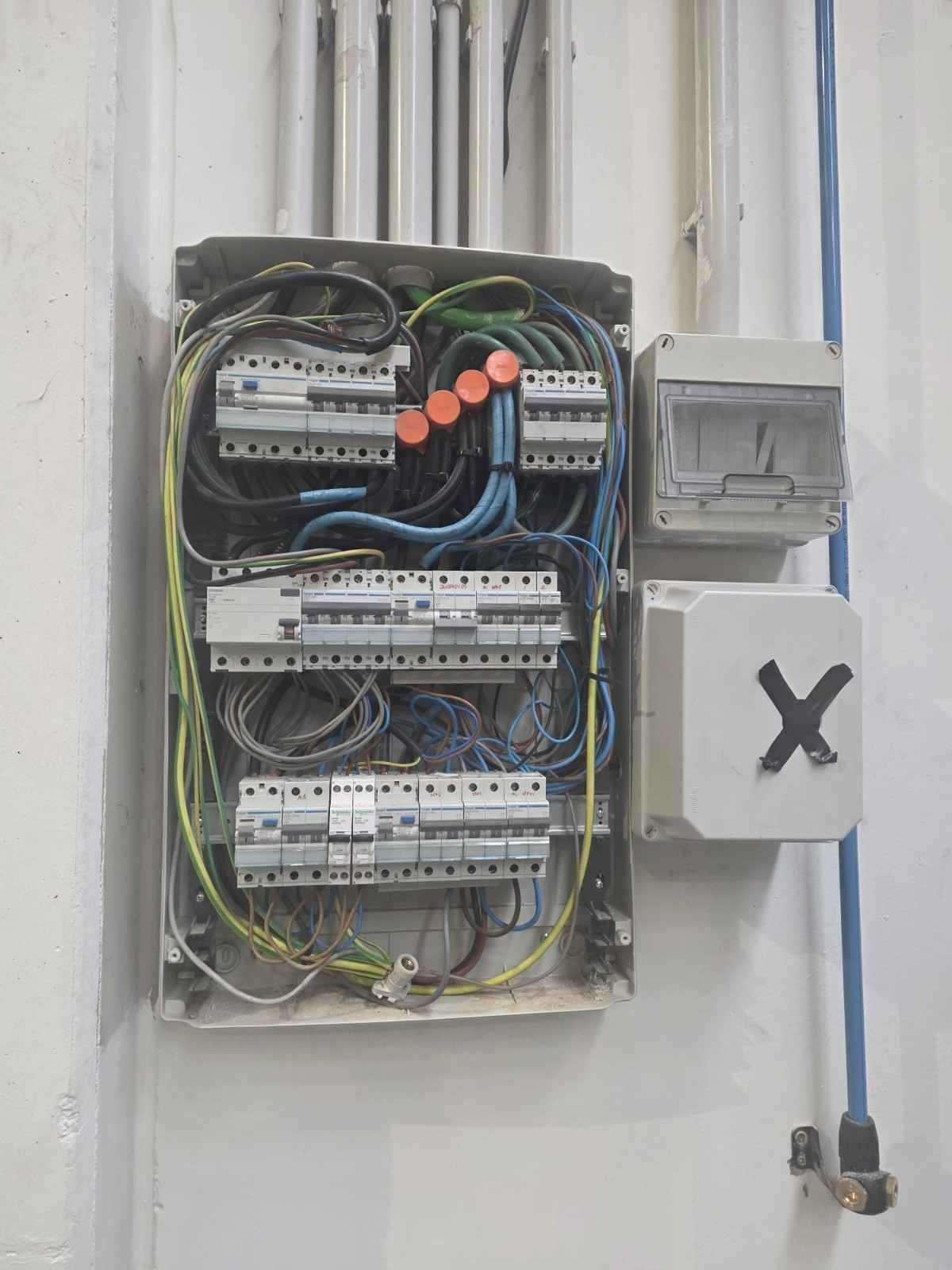 Panel eléctrico abierto con cables expuestos, montado en una pared blanca junto a una caja pequeña marcada con una X.
