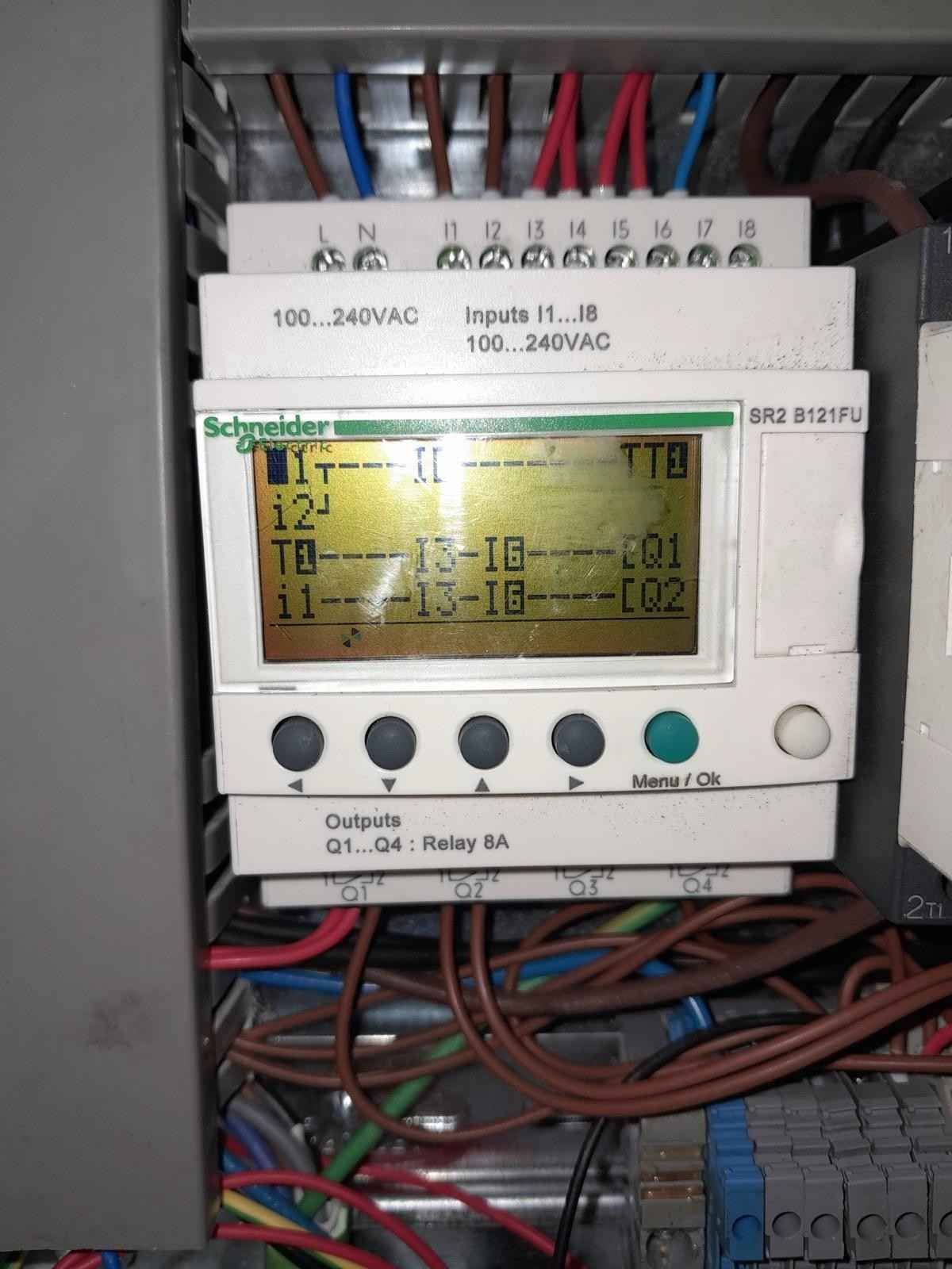 Panel de control junto a maquinaria industrial en una fábrica, con la puerta del armario abierta y equipos azules cerca