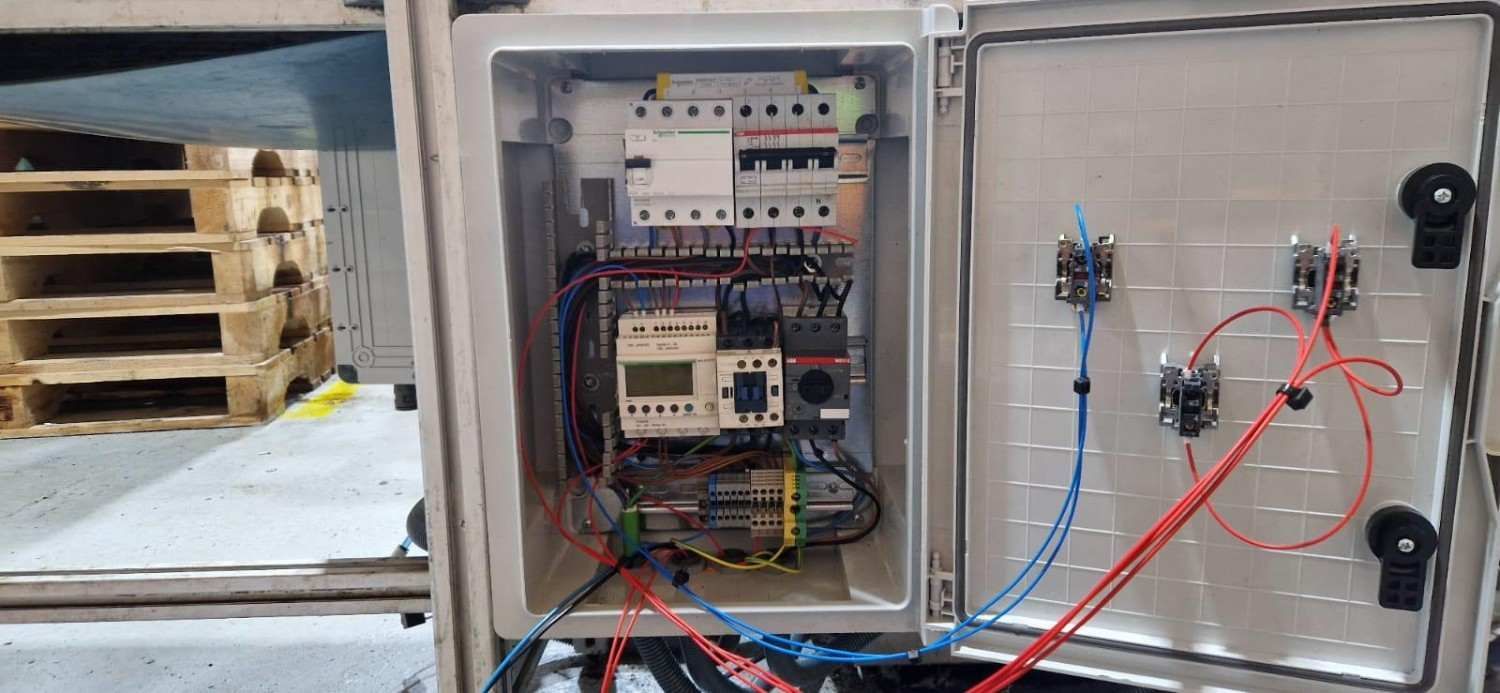 Panel eléctrico abierto con cables expuestos, montado en una pared blanca junto a una caja pequeña marcada con una X.