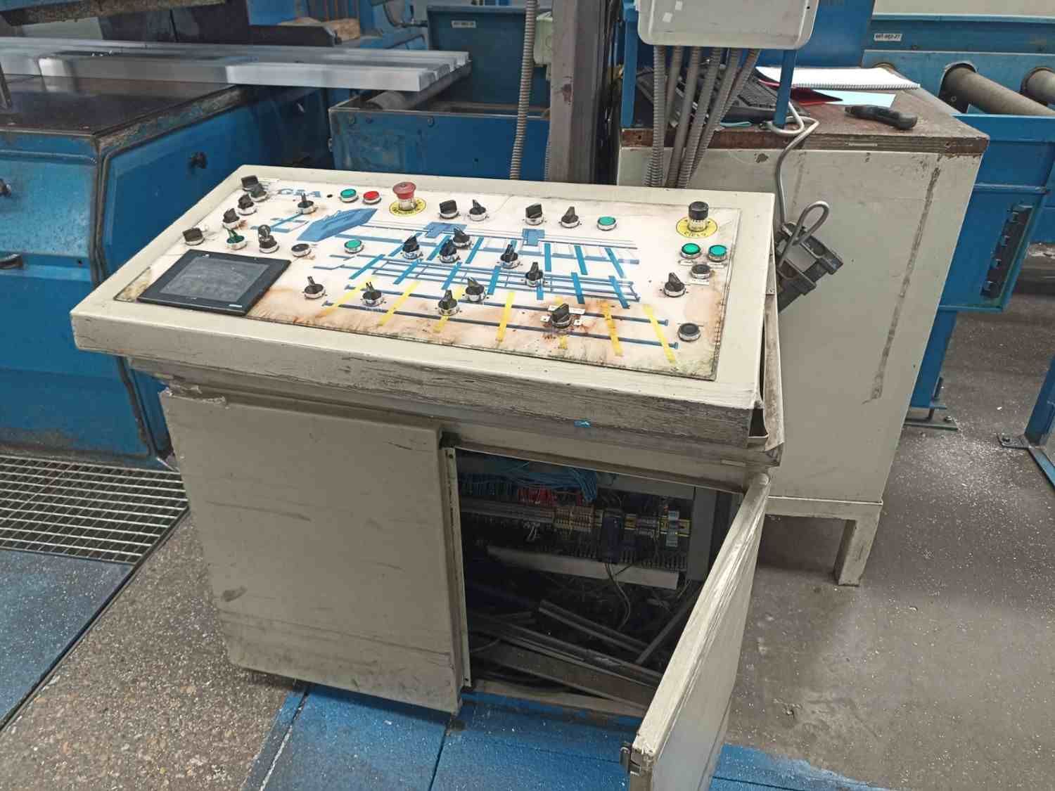 Panel de control junto a maquinaria industrial en una fábrica, con la puerta del armario abierta y equipos azules cerca