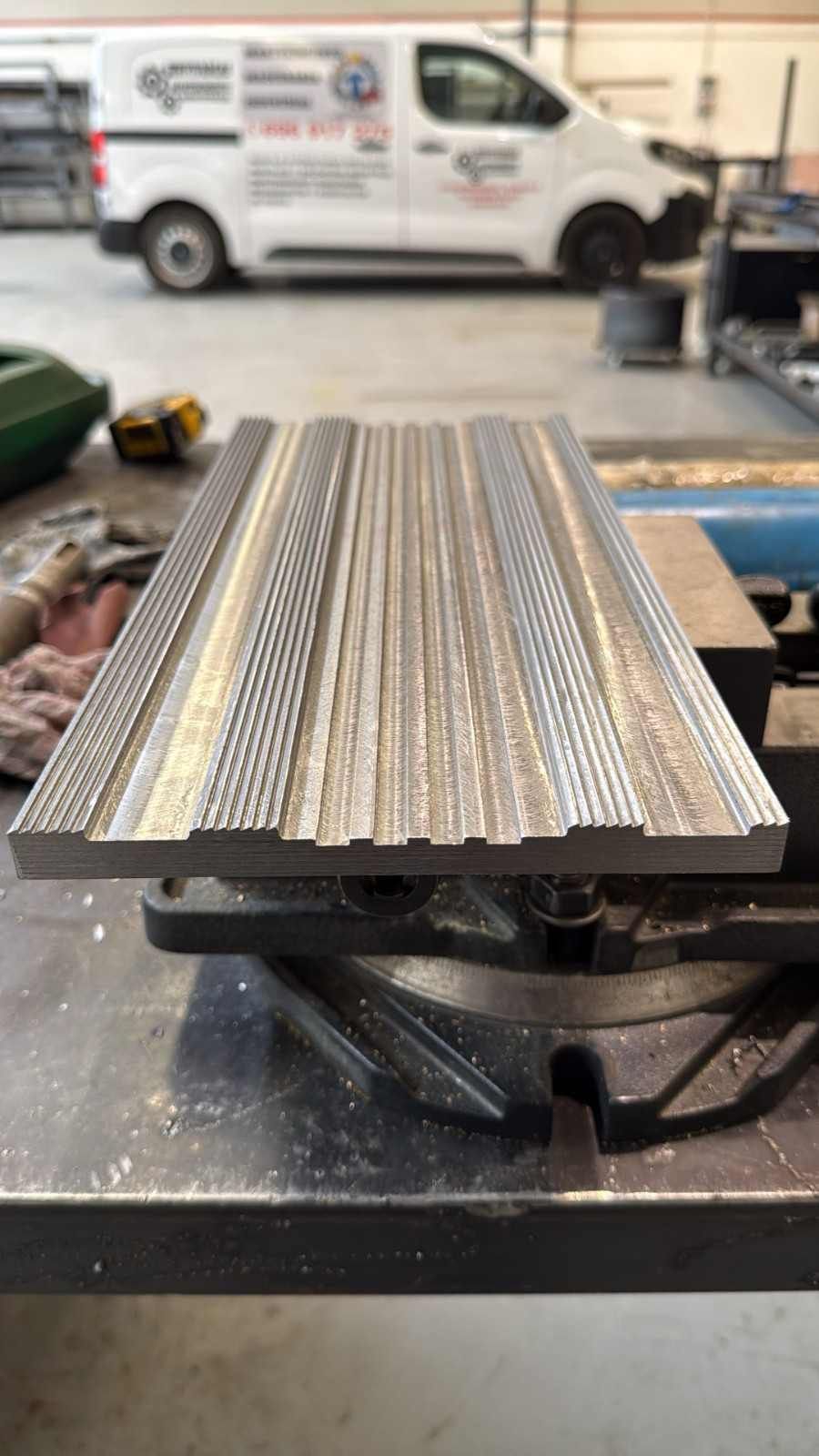 Tiras de metal con textura sujetas en un tornillo de banco de taller, con una furgoneta blanca desenfocada al fondo.