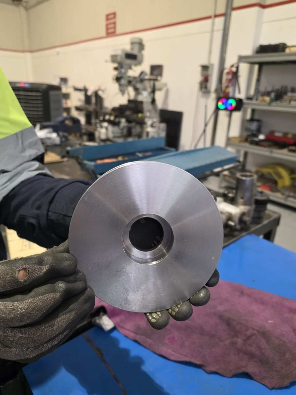 Manos enguantadas sosteniendo un disco de metal mecanizado en un taller, con herramientas y equipos al fondo