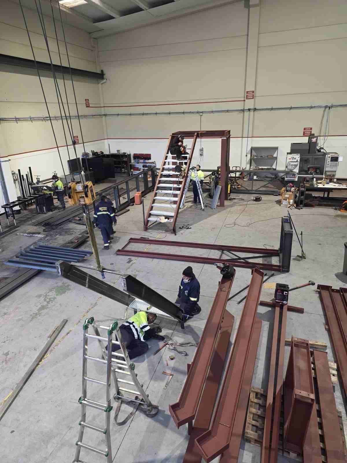 Taller industrial con trabajadores ensamblando una estructura de acero, escaleras, herramientas.