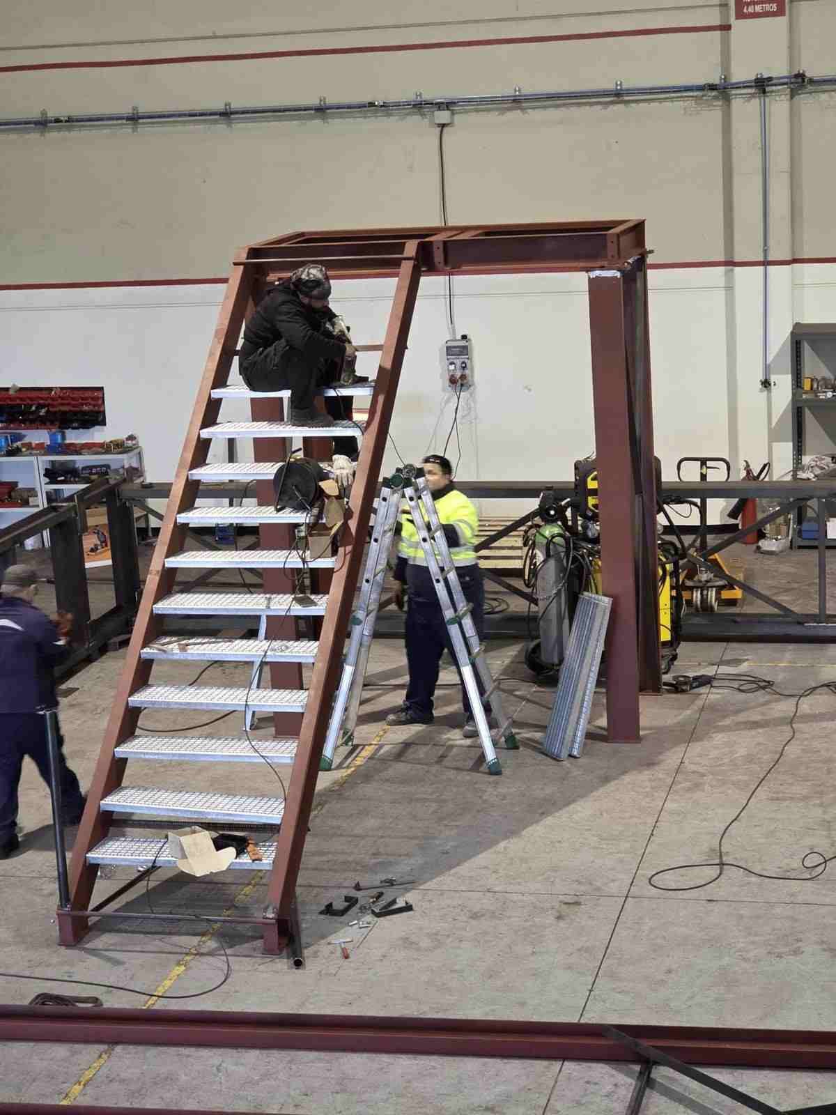 Trabajadores montando la estructura de una gran escalera metálica en un taller interior