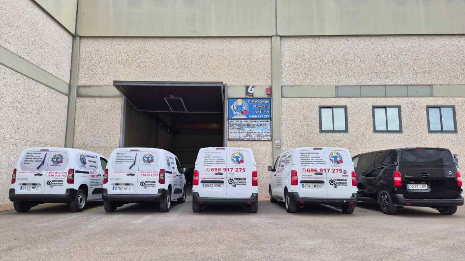 Cinco furgonetas blancas de servicio y un SUV negro aparcados frente a la zona de carga de un almacén.