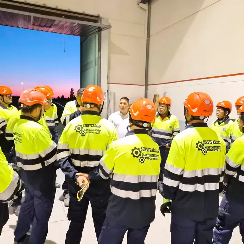 Grupo de trabajadores con equipo de seguridad y cascos dentro de un almacén, escuchando a un hombre.