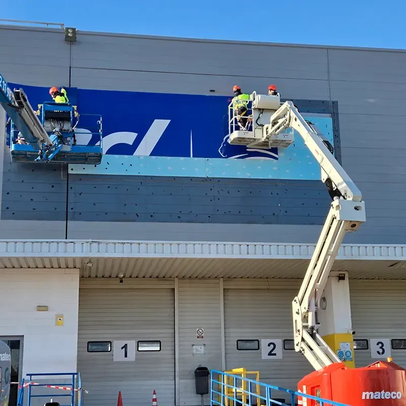 Trabajadores en ascensores instalando un logotipo azul y blanco en el exterior de un almacén con muelles de carga.