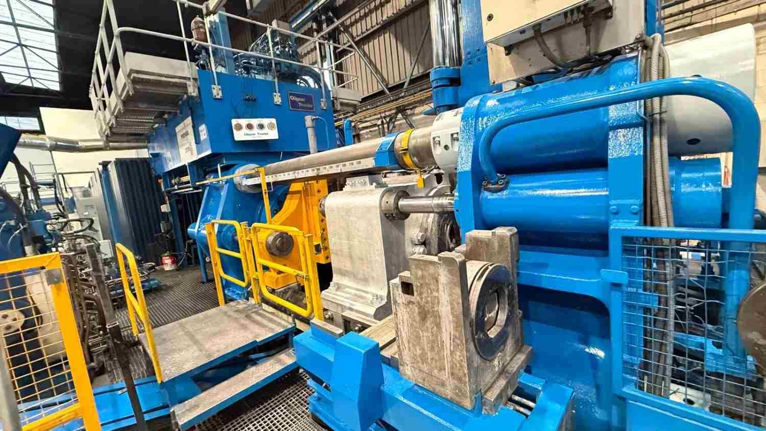 Maquinaria industrial azul en una fábrica, con barandillas de seguridad amarillas y tuberías metálicas.